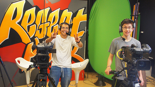 Two Youth Cabinet members wear headsets and stand behind video cameras.
