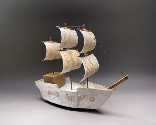 A detailed model of a ship with sails displayed against a neutral gray background, showcasing its intricate design.