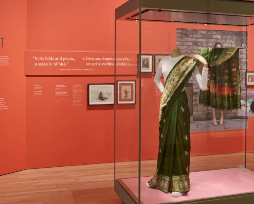Installation showing red text panels behind a glass case containing a green silk sari with woven metallic thread worn over a white T-shirt, shown on a mannequin wearing white running shoes.