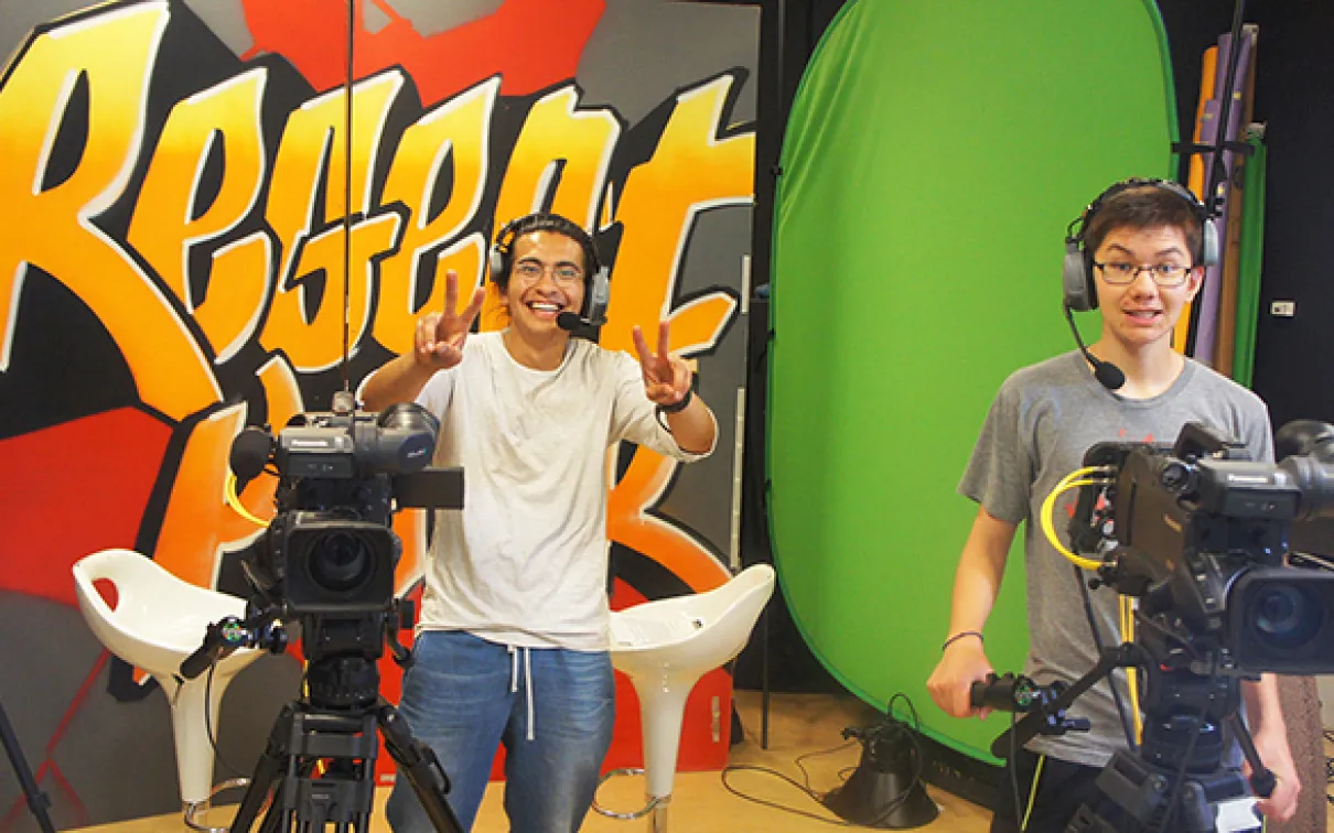 Two Youth Cabinet members wear headsets and stand behind video cameras.
